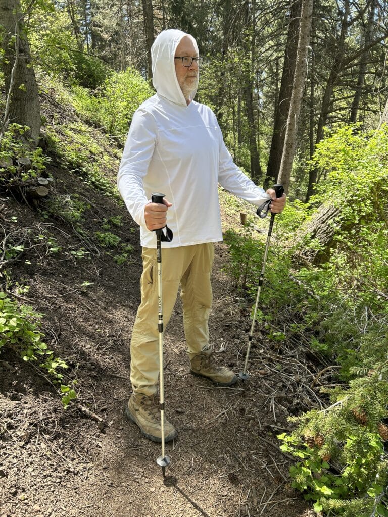 A man in a white hooed sun shirt and tan pants stands on a trail 