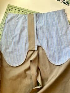 A pair of tan pants being sewn, with the inside front pockets showing. The pockets are blue striped cotton.
