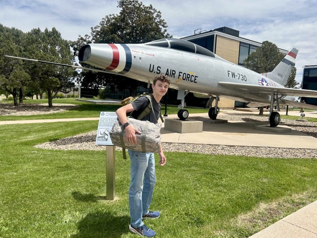 A tall teenage boy with a camo duffel bag stands in front of an aircraft display 