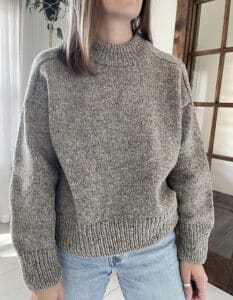A woman modeling a loose fitting chunky gray sweater. It has saddle shoulders and a high ribbed neck.