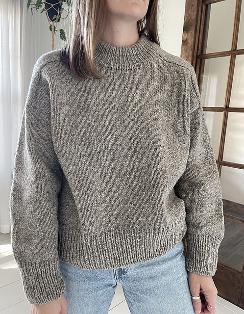 A woman modeling a loose fitting chunky gray sweater. It has saddle shoulders and a high ribbed neck.