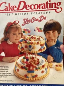 Cover of the 1987 Wilton Cake Decorating Yearbook, with a boy and girl gazing at a tiered cake decorated with clowns and balloons.