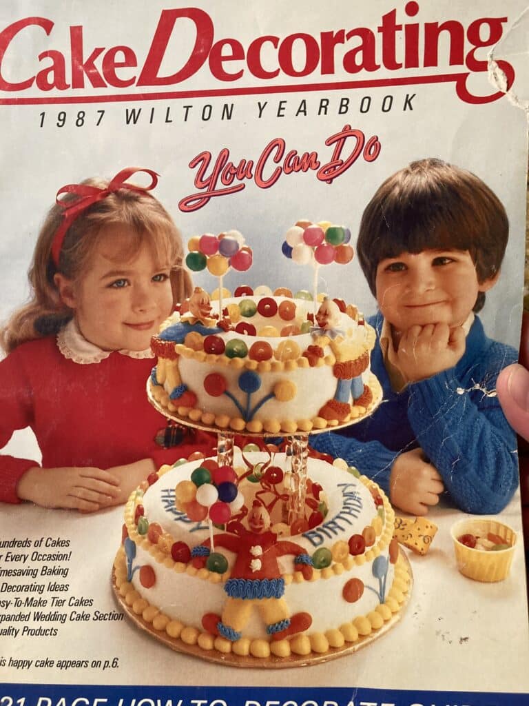 Cover of the 1987 Wilton Cake Decorating Yearbook, with a boy and girl gazing at a tiered cake decorated with clowns and balloons.