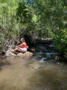 A woman sitting on a rock in a river. She's wearing short shorts , a tank top, and a baseball hat.