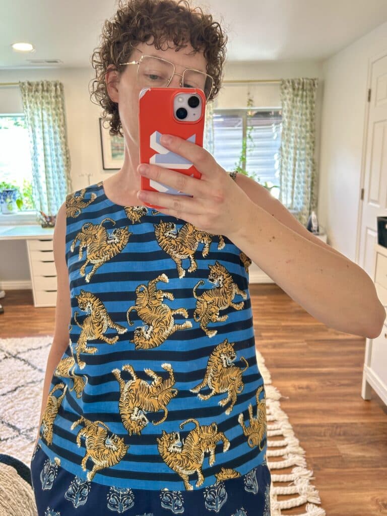 A woman takes a mirror selfie. She's wearing a sleeveless top with horizontal stripes in bright blue and navy blue, with Tibetan style tigers scattered over the stripes. 