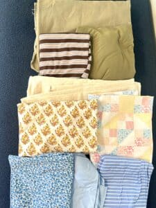 A pile of nine fabrics in olive, creams, and blues, organized in rows by color