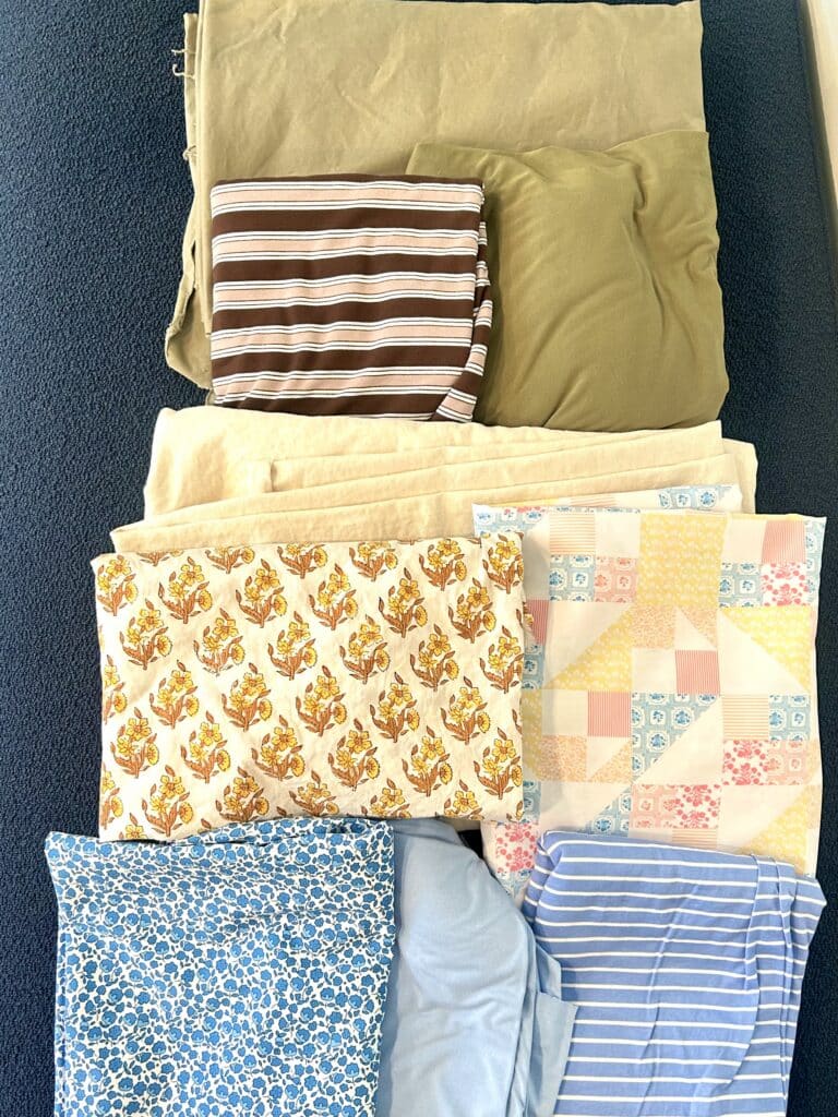 A pile of nine fabrics in olive, creams, and blues, organized in rows by color