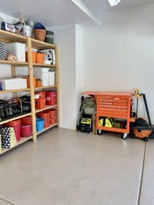 The same corner of the garage but with smooth painted white walls and a tan speckeld epoxy floor. The empty shelves are filled with tidy boxes and there is an orange tool chest against the wall.