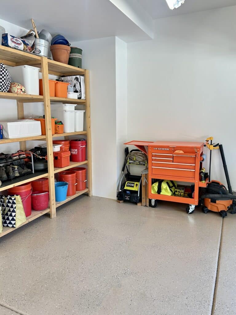 The same corner of the garage but with smooth painted white walls and a tan speckeld epoxy floor. The empty shelves are filled with tidy boxes and there is an orange tool chest against the wall. 