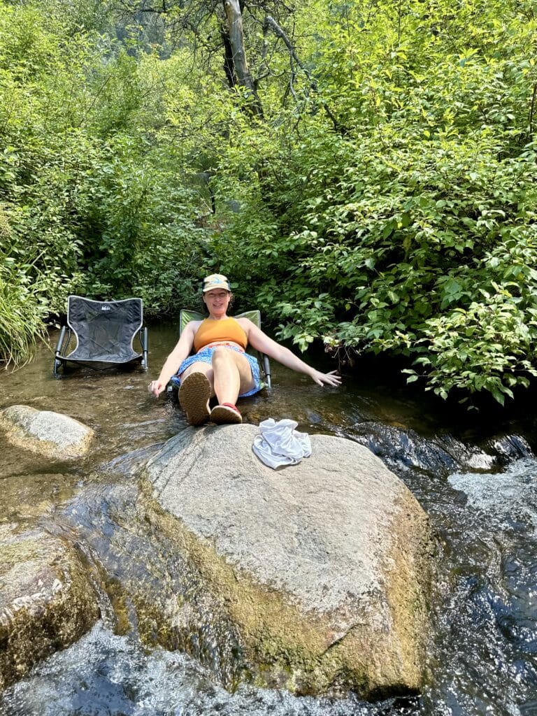 A woman sitting on a low camp chair in shallow part of a stream. Her feet are up on a big rock that the water is cascading over. Trees are in the background. 