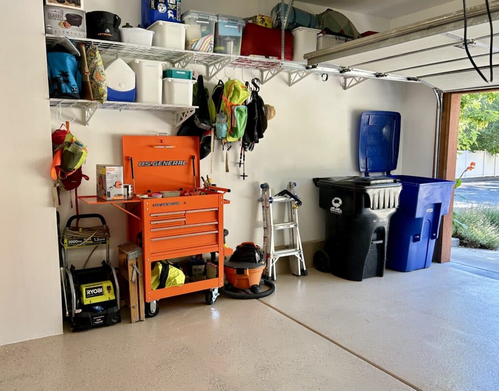 The left wall of a garage with white walls and beige floors. There is a line of high wire shelving at the ceiling. On the floor below are a pressure washer, orange tool chest, ladder, and two garbage cans.