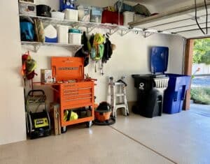 The left wall of a garage with white walls and beige floors. There is a line of high wire shelving at the ceiling. On the floor below are a pressure washer, orange tool chest, ladder, and two garbage cans.