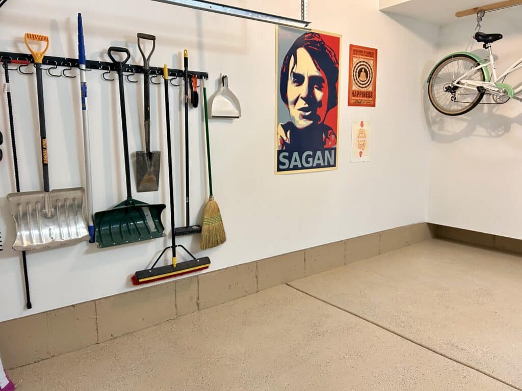 The right wall of a garage with white walls and a tan floor. There is a line of hanging yard tools and a large poster of Carl Sagan and two smaller posters.