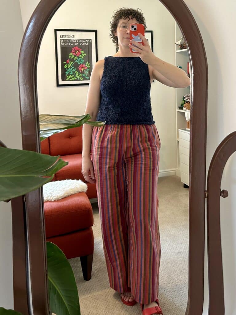 A woman wearing burgunfy stripe pants and a sleeveless boatneck navy knitted top takes a mirror selfie