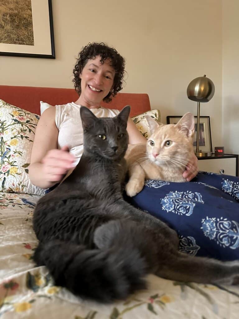 A grinning woman on a bed with a gray cat and an orange cat on her lap