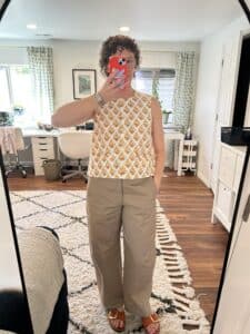 A woman takes a mirror selfie. She's wearing a sleeveless yellow-on-white block print vest and tan balloon pants