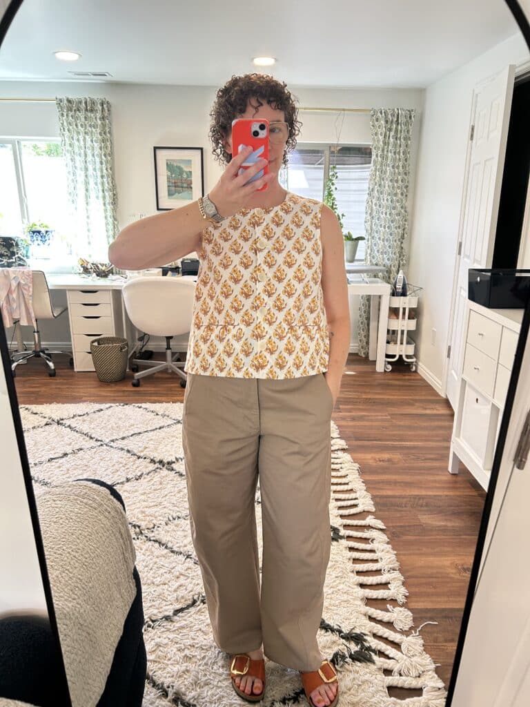 A woman takes a mirror selfie. She's wearing a sleeveless yellow-on-white block print vest and tan balloon pants