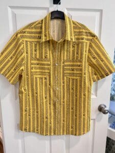 A shirt on a hanger. The background is yellow with gray tire and track lines and a CAT Logo. There are pearl snaps and a curved yoke.