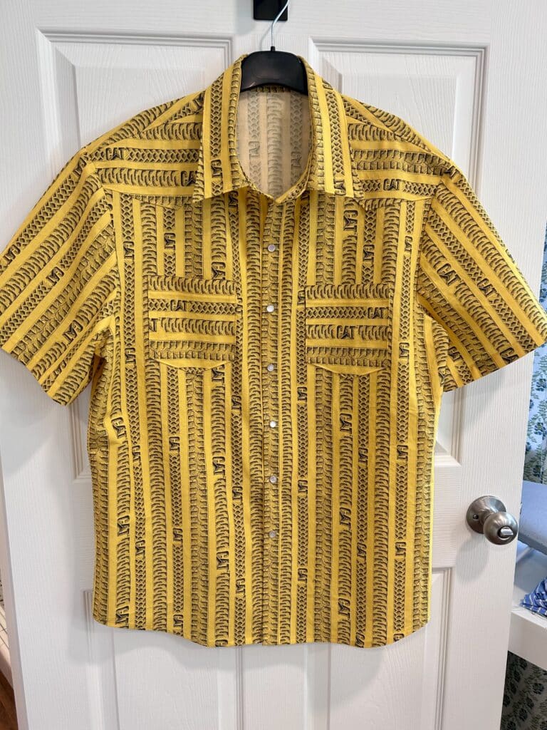 A shirt on a hanger. The background is yellow with gray tire and track lines and a CAT Logo. There are pearl snaps and a curved yoke.