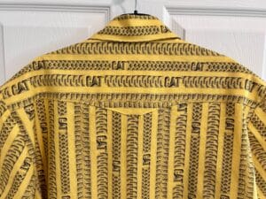 Detail of the back yoke of a shirt. The fabric is yellow with gray tire and track marks on it, with the CAT logo.