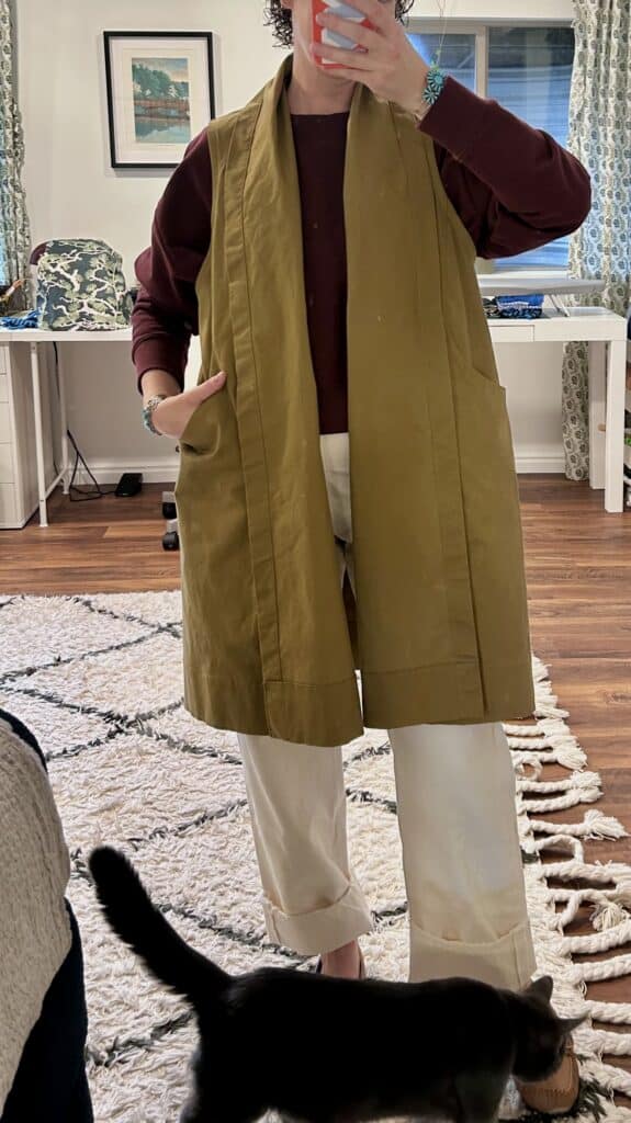 A woman takes a mirror selfie with a gray cat by her feet. She's wearing a long tan vest with pockets