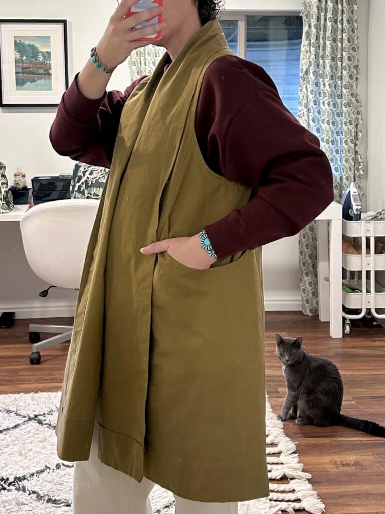 A woman takes a mirror selfie in a long tan vest with pockets. The vest is knee length and voluminous. 