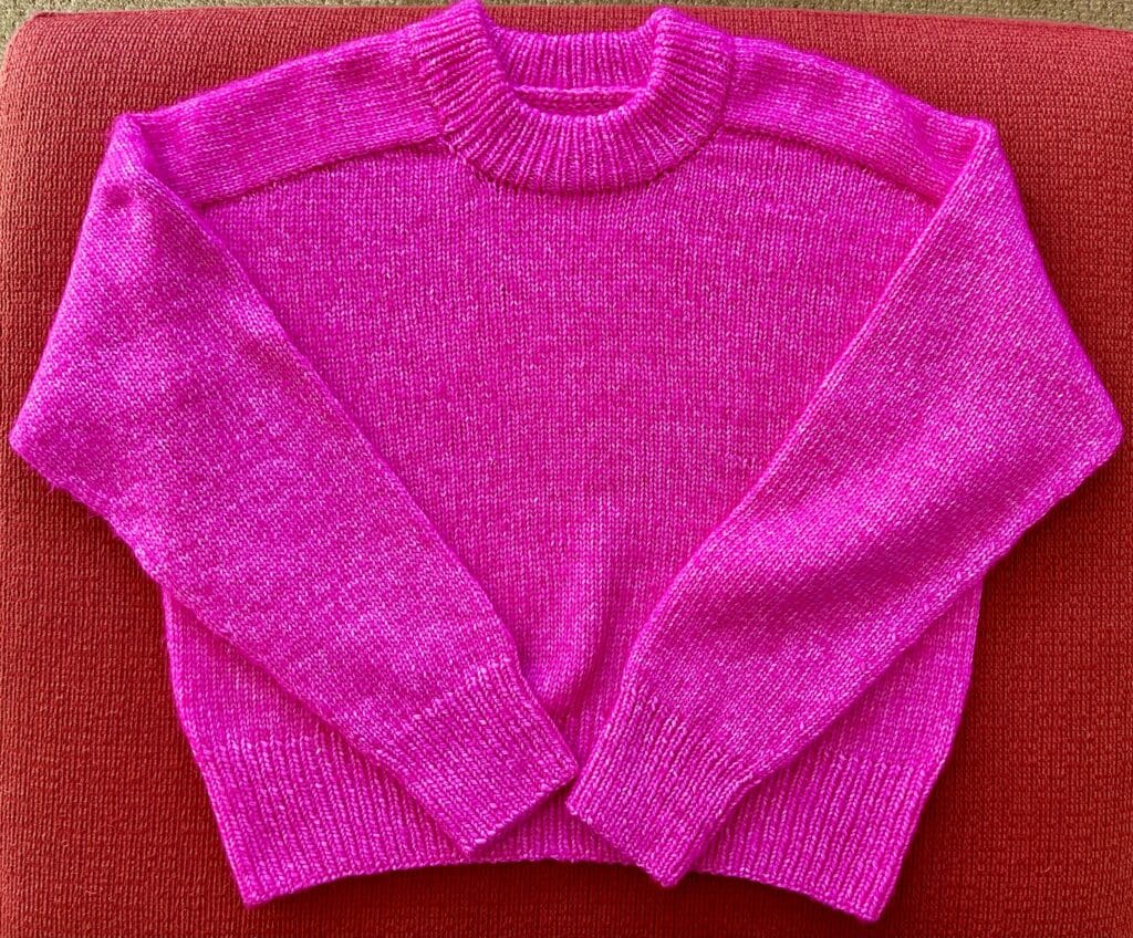 A bright pink saddle shoulder sweater laid flat on an orange background