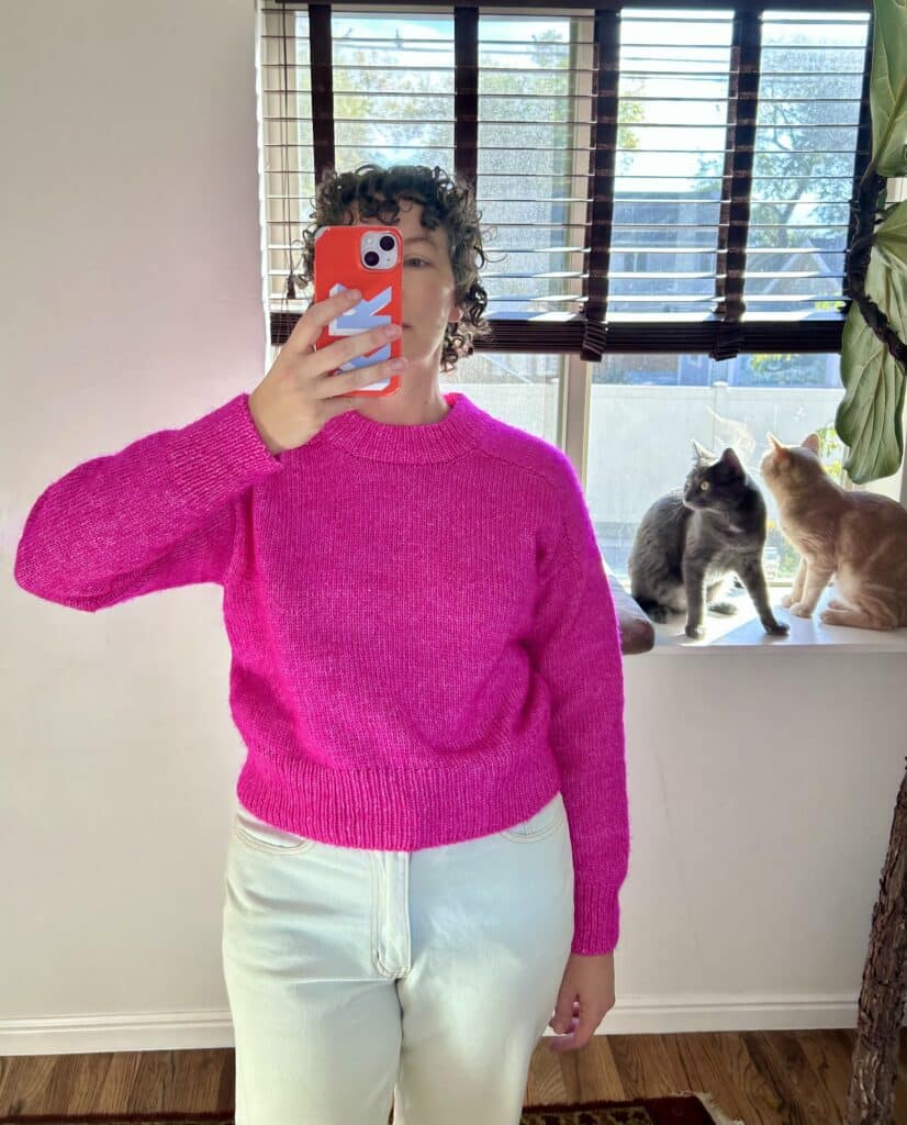 A woman in a bright pink sweater and light wash jeans takes a selfie in front of a mirror. There are two cats in the background. 